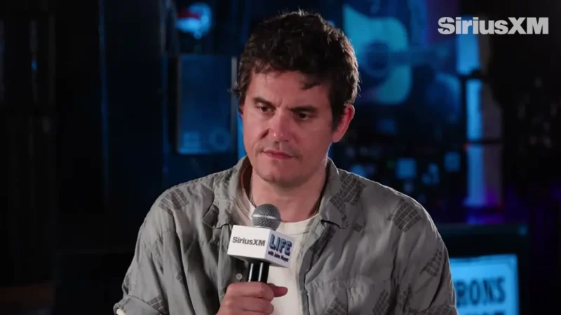 John Mayer in a gray short-sleeve shirt holding a microphone during a SiriusXM interview