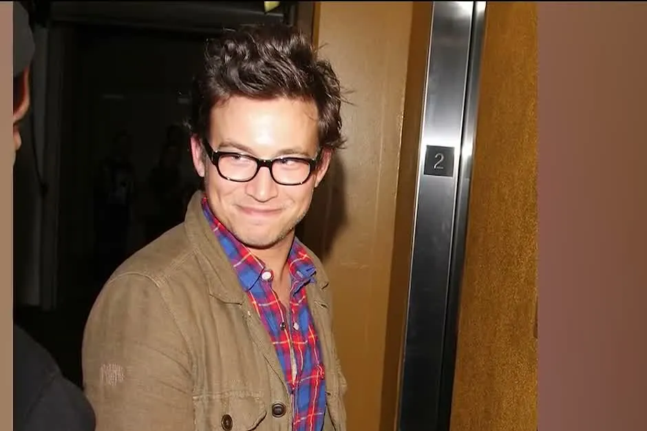 Jonathan Taylor Thomas as an adult, in dark-rimmed glasses and a tan jacket over a plaid shirt