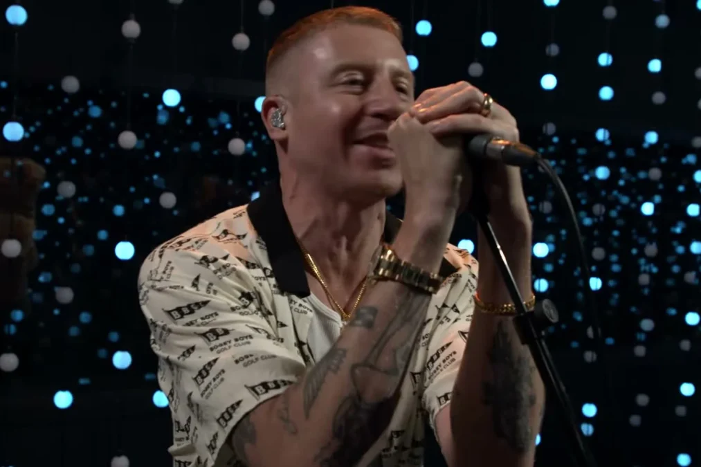 Macklemore performing into a handheld microphone under stage lights
