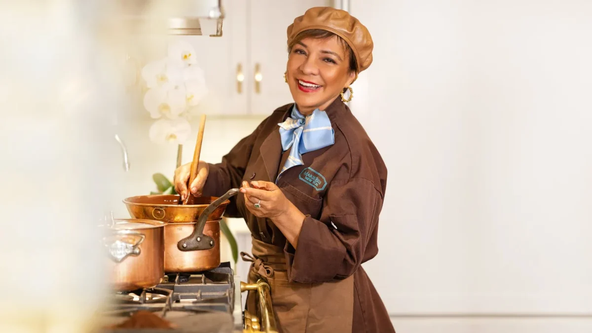 Maribel Lieberman, founder and CEO of MarieBelle New York, smiling in a brown chef's coat and beret as she stirs chocolate in a copper pot
