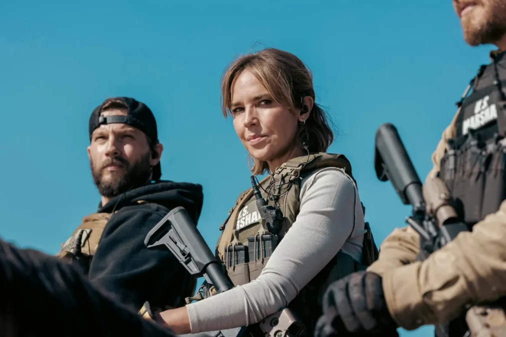 Arielle Kebbel as Marshal Belle Skinner in a tactical vest, holding a rifle beside two armed teammates under a blue sky