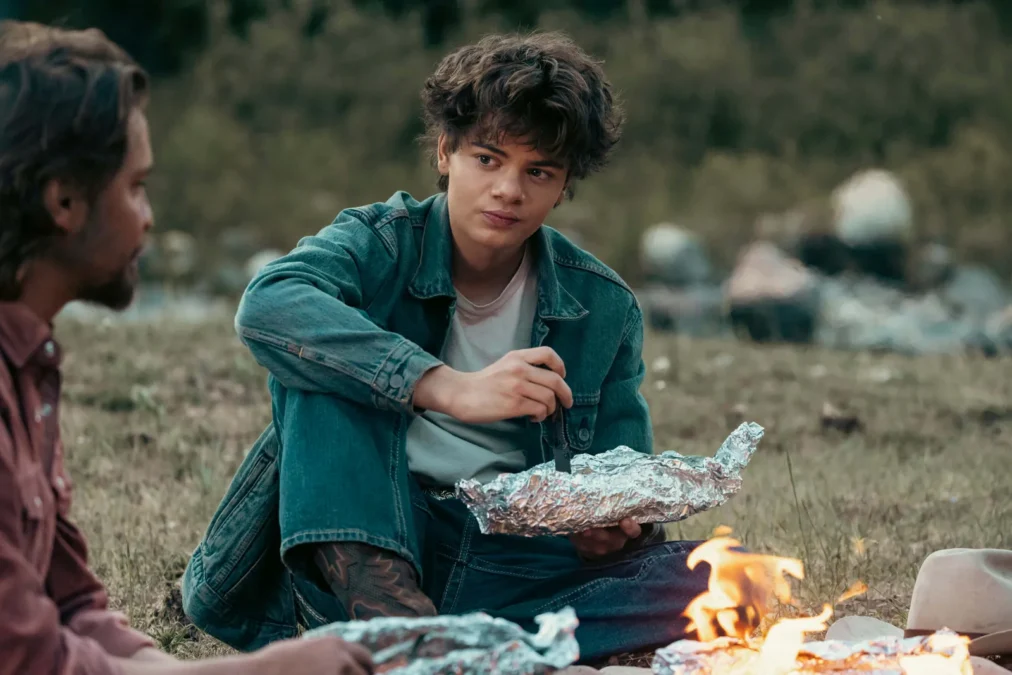 Brecken Merrill as Tate Dutton sits by a campfire in a denim jacket, eating from foil, with another man beside him