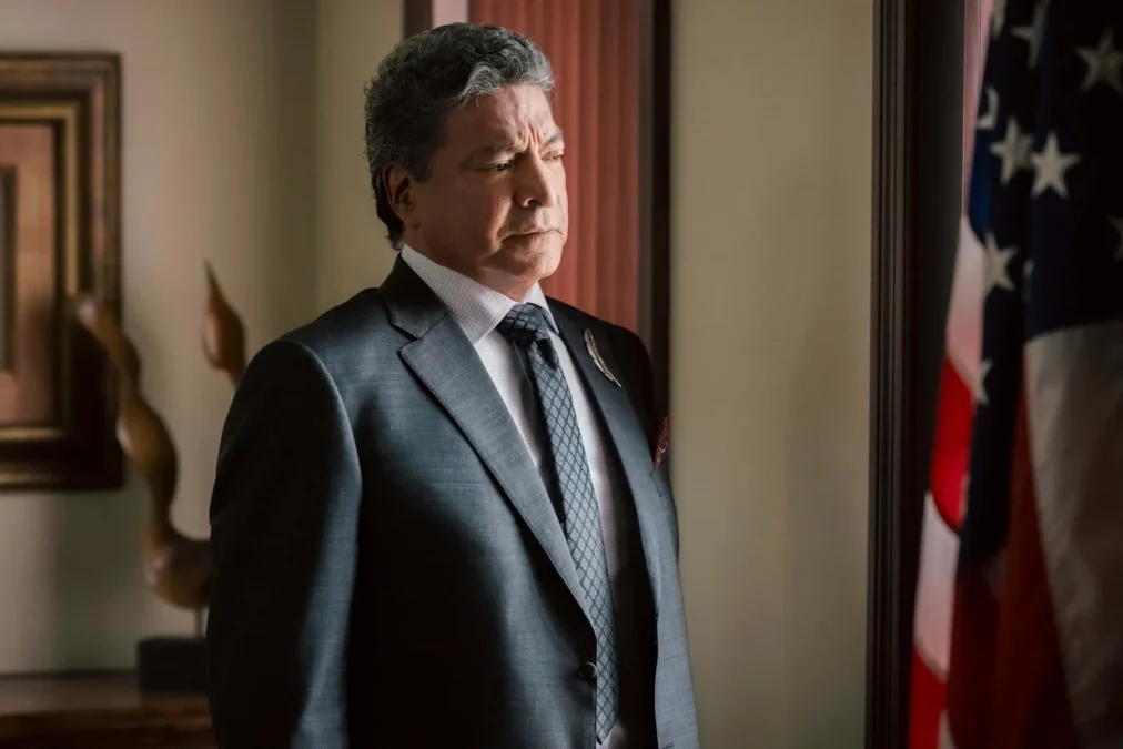 Gil Birmingham as Thomas Rainwater in a gray suit, standing in an office beside an American flag