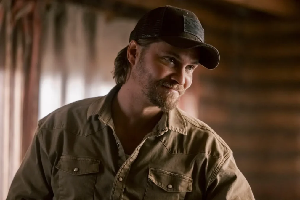 Luke Grimes as Kayce Dutton in a ball cap and tan work shirt, smiling inside a wood-paneled room