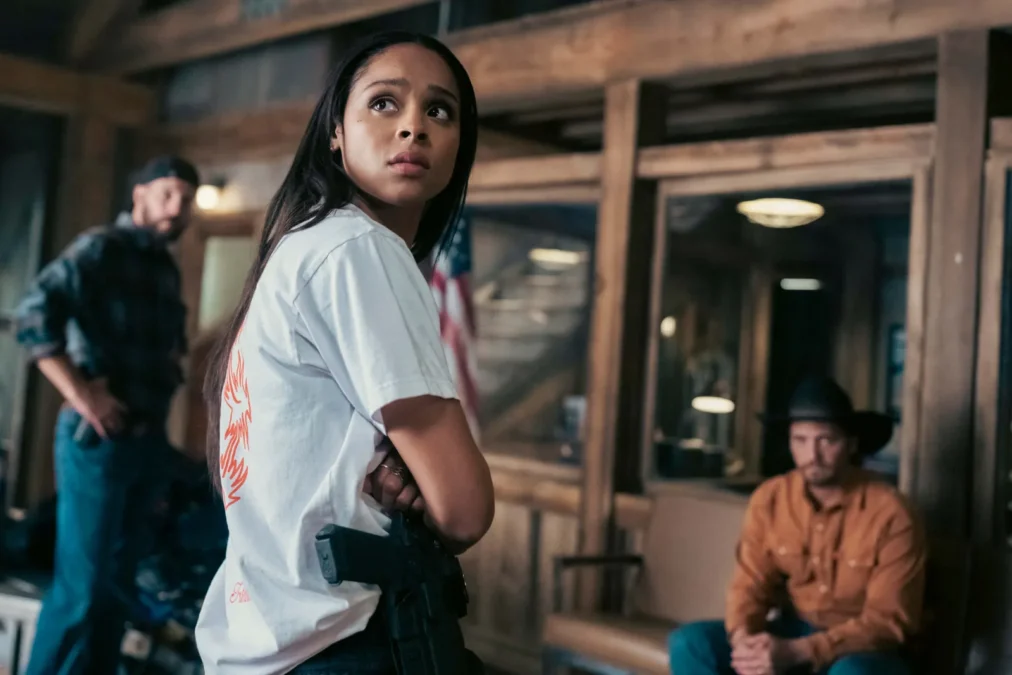 Ash Santos as Andrea Cruz in a white T-shirt with a holstered gun, with Kayce Dutton seated in the background