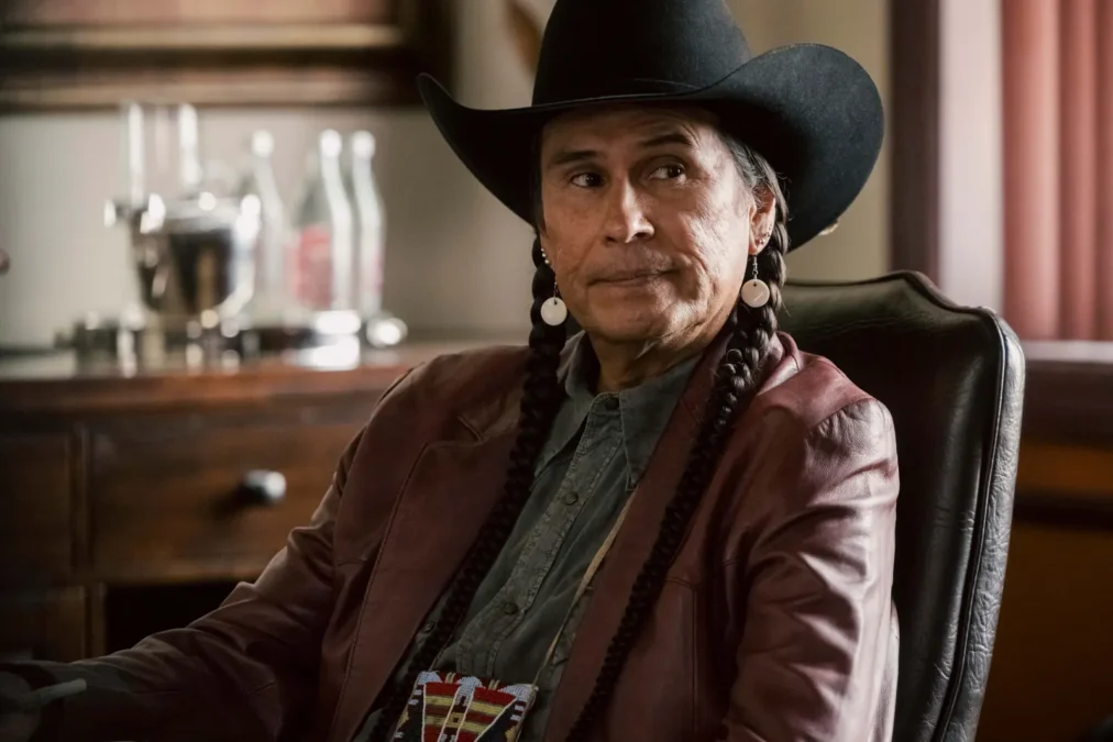 Mo Brings Plenty as Mo in a black cowboy hat and braids, seated in a leather chair