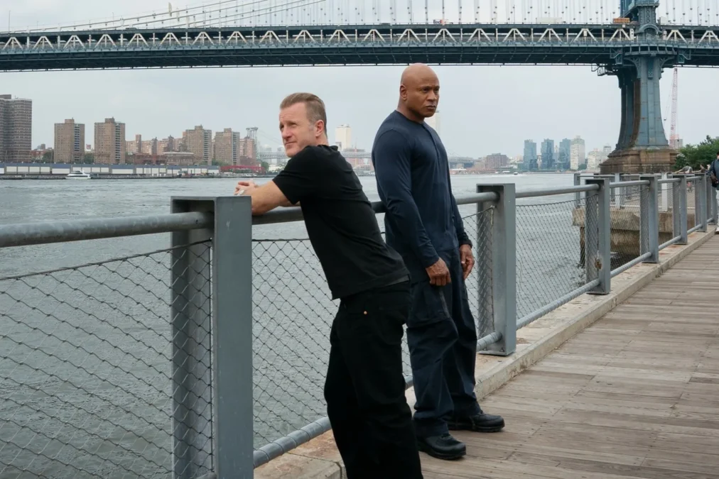 Scott Caan in a black T-shirt leaning on a railing beside LL COOL J on a waterfront boardwalk under a suspension bridge