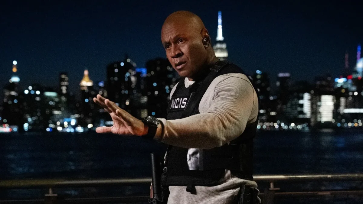 LL COOL J as Sam Hanna in an NCIS vest at night, gesturing with one hand, with the lit Manhattan skyline behind him
