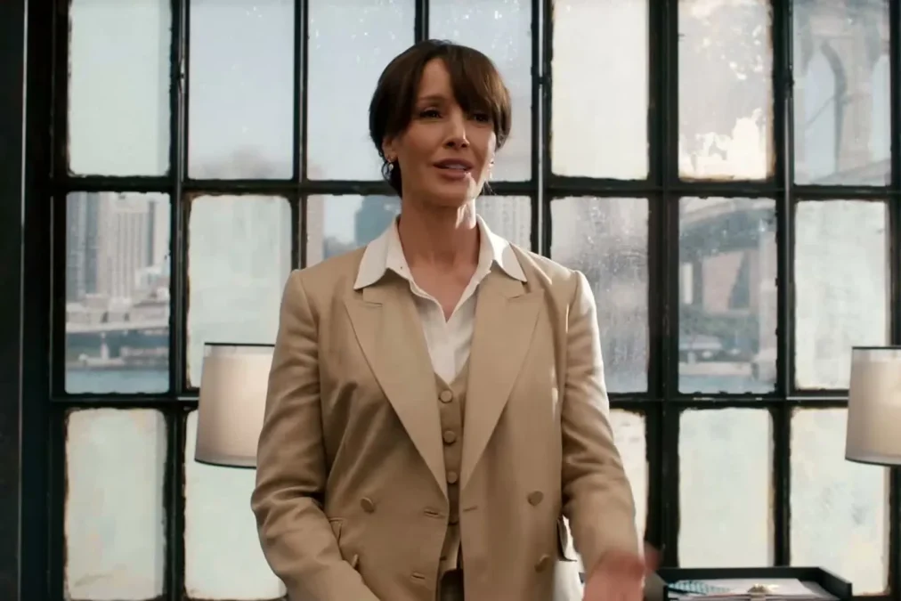 Jennifer Beals in a beige suit standing in front of tall factory-style windows