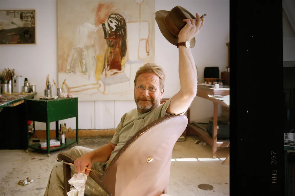 Martin Mull raises a hat while sitting in his painting studio among canvases and art supplies