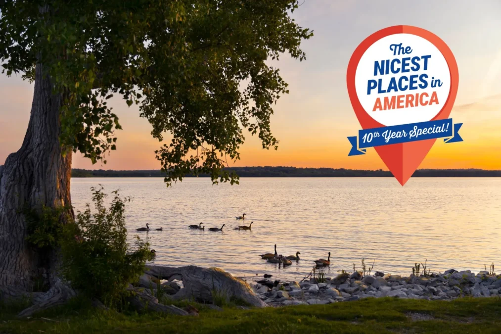 A lake at sunset with Canada geese swimming near a rocky shore under a large tree, with a Nicest Places in America 10th Year Special badge