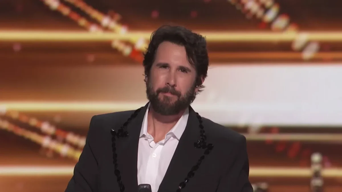 Josh Groban, bearded, in a black jacket with beaded lapels and a white shirt on the America's Got Talent stage