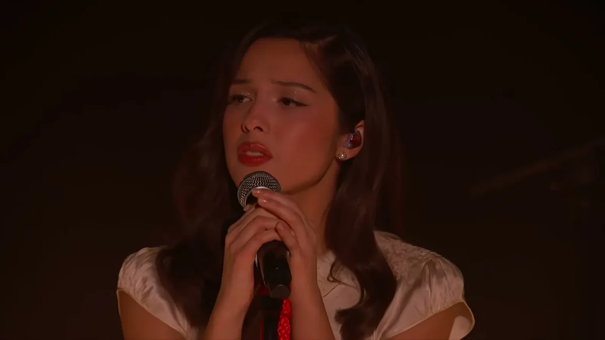 Olivia Rodrigo singing into a handheld microphone under red stage lights in a cream dress