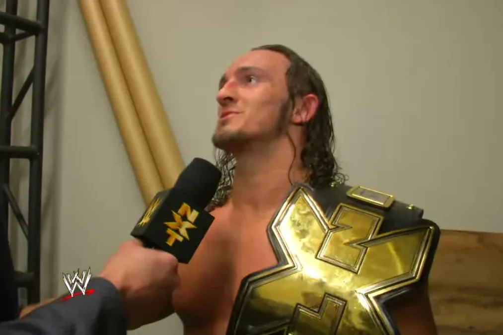 Adrian Neville backstage with the gold NXT Championship belt over his shoulder, speaking into an NXT microphone