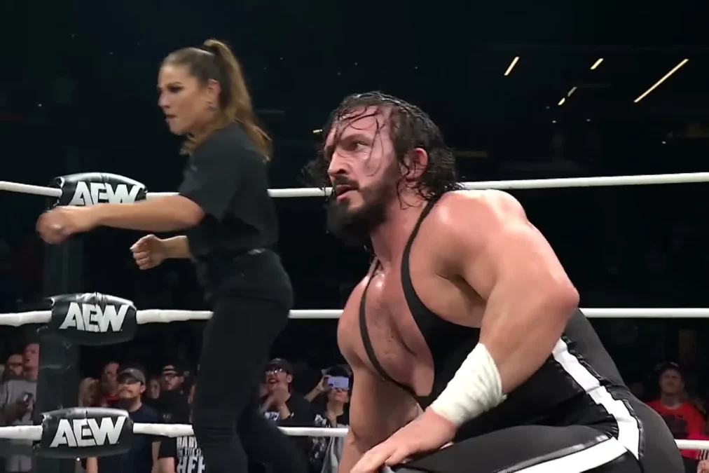 PAC in a black singlet crouching at ringside on AEW TV, with a woman in black behind him at the ring apron