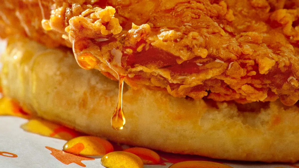 Close-up of honey sauce dripping from a crispy fried chicken breast onto a buttermilk biscuit on Popeyes paper