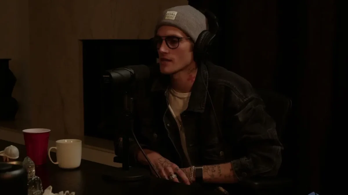 Presley Gerber seated at a podcast table in a denim jacket, with tattoos on his neck and hands
