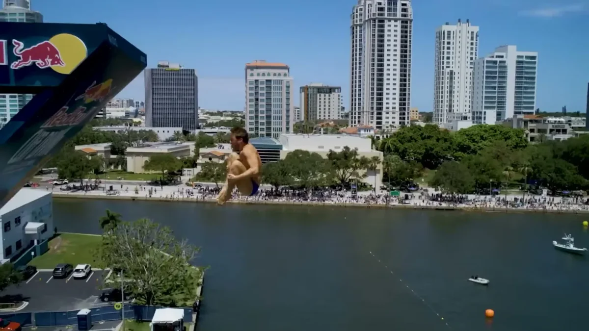 A cliff diver tucked in midair beside a Red Bull platform above the water at St. Pete Pier, with crowds on the shore and downtown St. Petersburg towers behind
