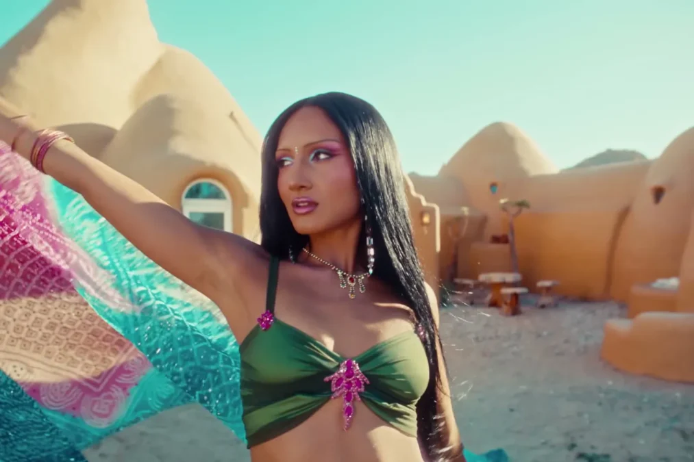 Rhea Raj in a green top and jewelry, one arm raised with a flowing scarf, in front of domed desert buildings in the Mumbai music video