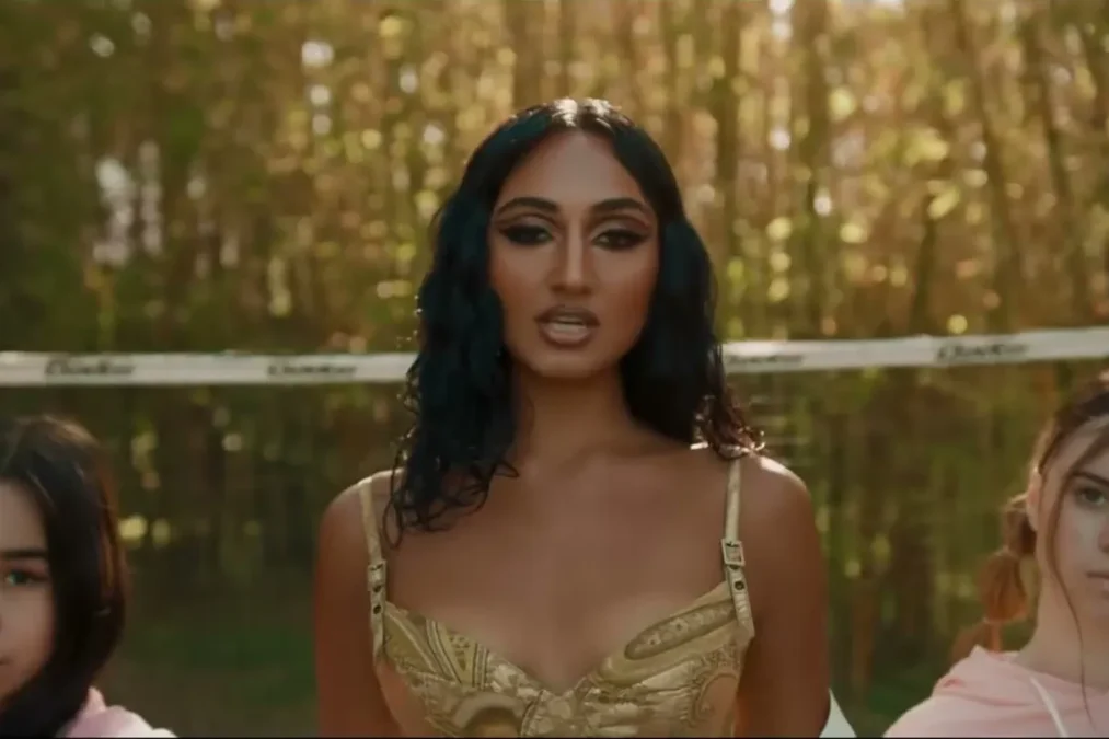 Rhea Raj in a gold corset top, standing between two young dancers in front of a bamboo fence in the Outside music video