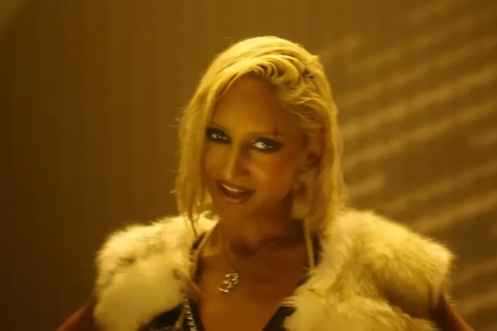 Rhea Raj in a platinum blond wig and a white fur jacket under warm light in the HUSH music video