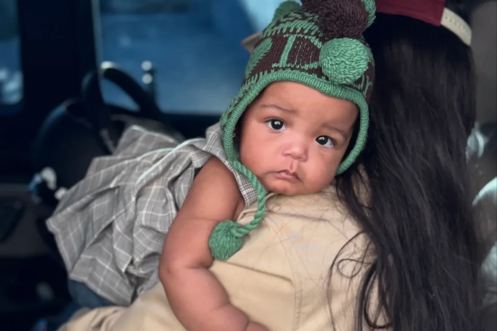 Baby Rocki in a green knit hat with pom-poms, held over the shoulder of a woman with long dark hair