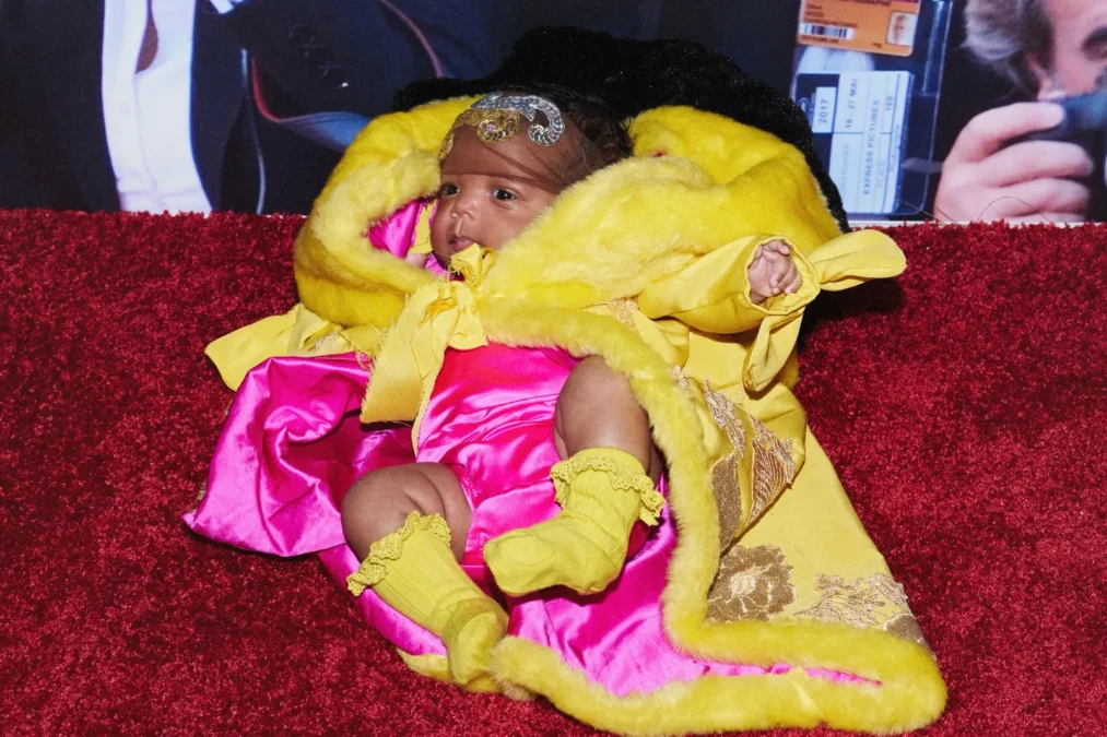 Baby Rocki on a red carpet in a yellow fur-trimmed cape with gold embroidery, in front of a backdrop of photographers