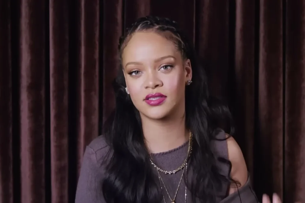 Rihanna with braided hair and pink lipstick talking in front of a dark velvet curtain in a Vogue video