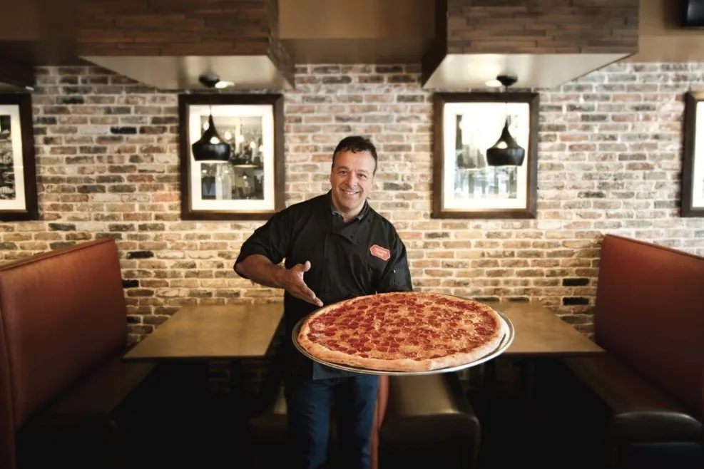 Chef Anthony Russo, founder of Russo's New York Pizzeria, holding a large pepperoni pizza in a restaurant dining room with brick walls