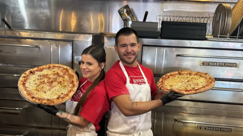 Kristina and Christian Alvarez, owners of the Russo's New York Pizzeria in Tempe, Arizona, in red T-shirts and white aprons, holding New York-style cheese pizzas in front of the pizza ovens