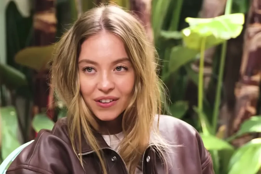 Sydney Sweeney in a brown leather jacket smiling during a GQ interview in a garden