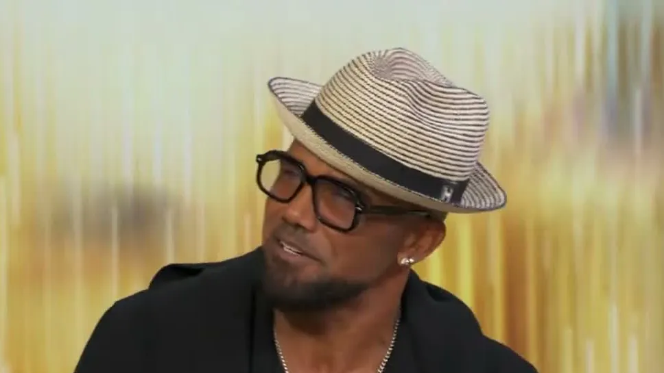 Shemar Moore in a straw fedora and dark-rimmed glasses during a TV interview