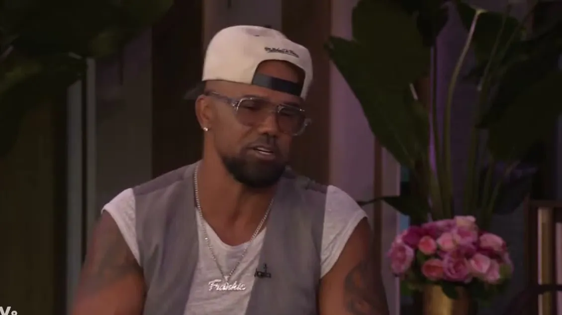 Shemar Moore in a white cap, clear glasses and a gray vest talking on a TV talk show set
