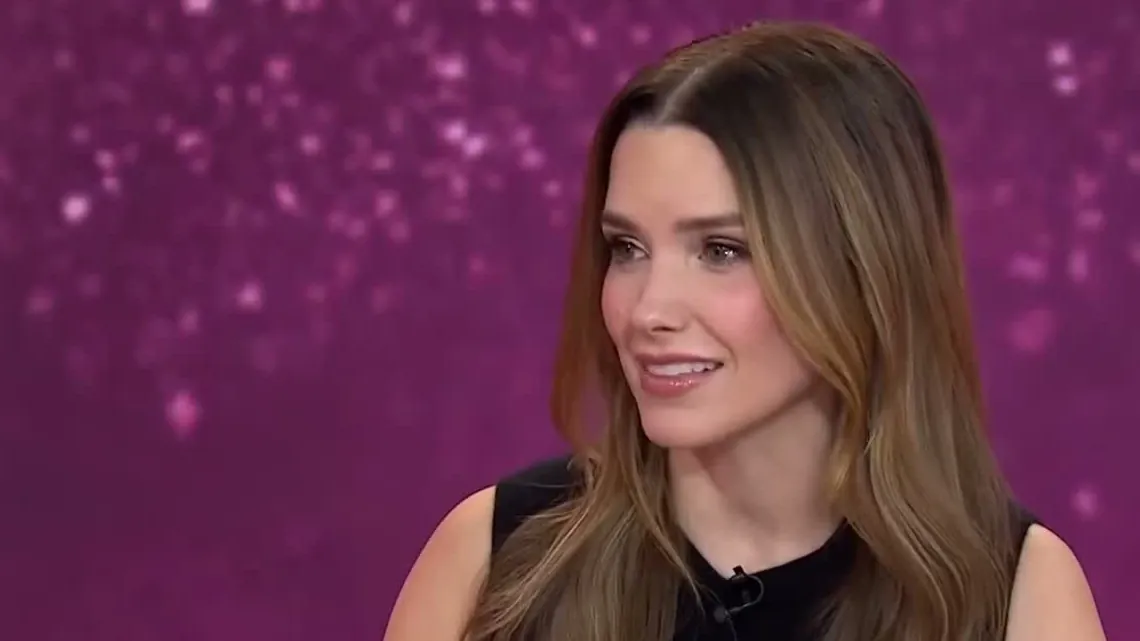 Sophia Bush with long brown hair in a black sleeveless top smiling on a talk show set