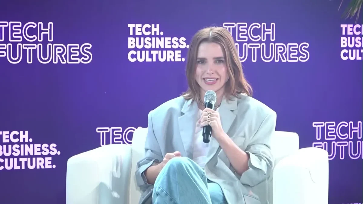 Sophia Bush in a light gray blazer holding a microphone on a stage with a purple backdrop