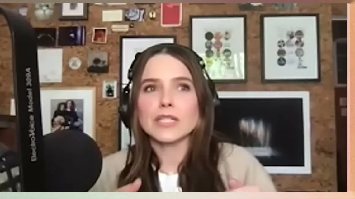 Sophia Bush wearing headphones talking on a video call in front of a cork board with framed photos