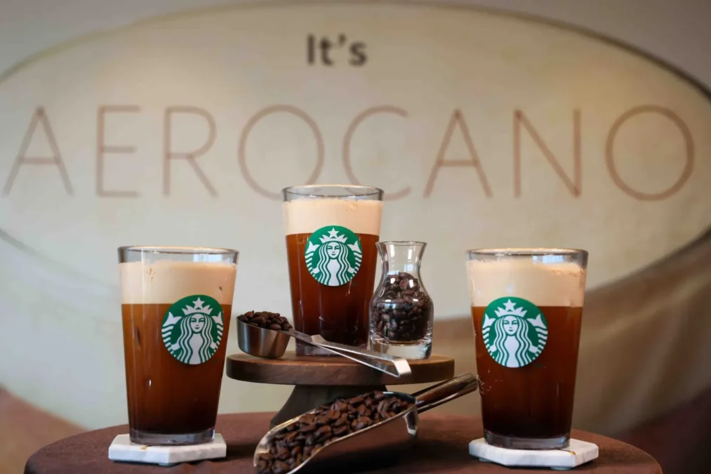 Three Starbucks Aerocano drinks in glasses with foam tops, beside a jar of coffee beans, in front of a sign reading 'It's Aerocano' at the drink's South Korean launch