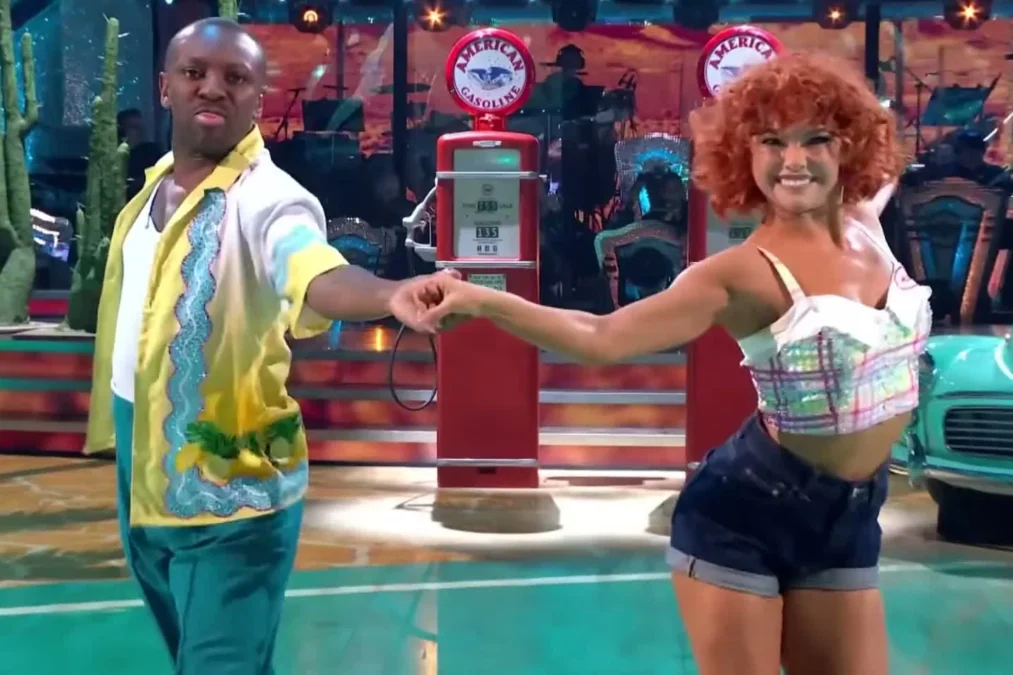 Shaun Wright-Phillips in a yellow and green shirt holding hands with red-haired pro dancer Alexis Warr on a gas-station set on Strictly Come Dancing