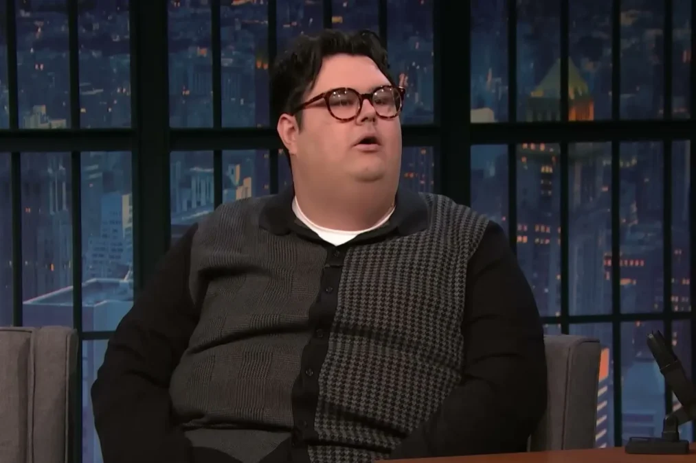 Caleb Hearon in glasses and a gray houndstooth cardigan sitting on the Late Night with Seth Meyers couch