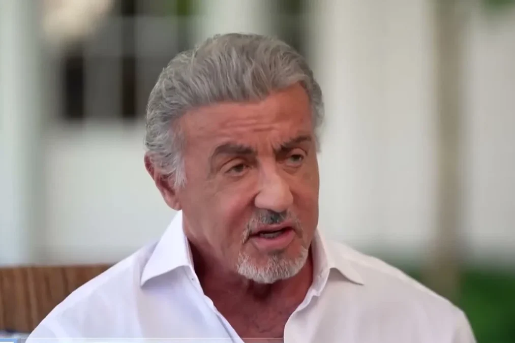 Sylvester Stallone in a white open-collar shirt talking during an outdoor TV interview
