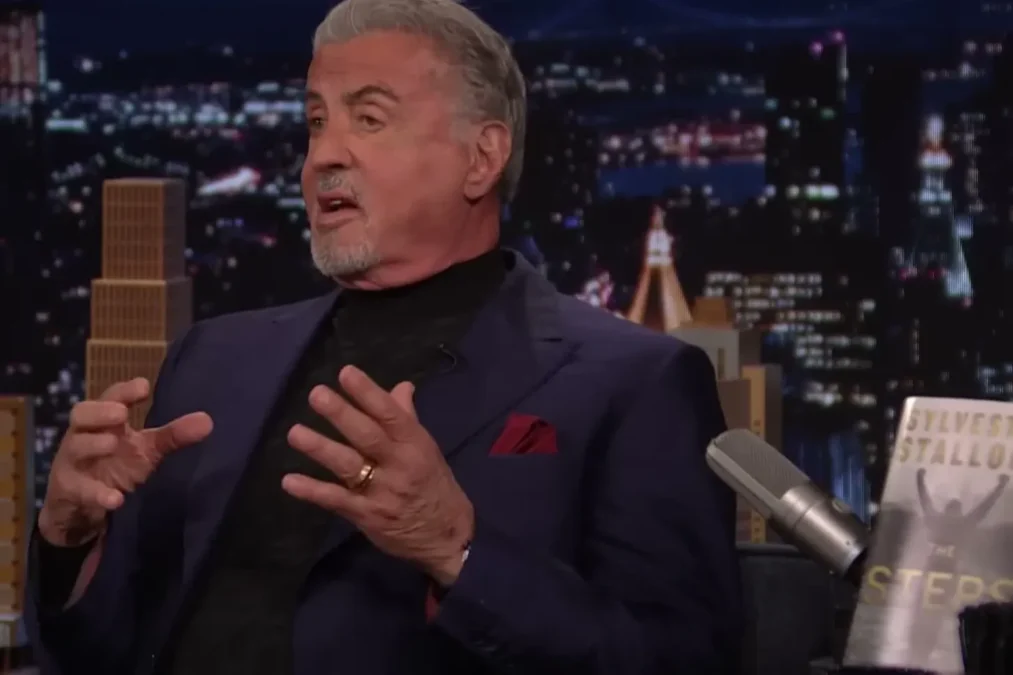 Sylvester Stallone gesturing on The Tonight Show set, with a copy of his memoir The Steps on the desk
