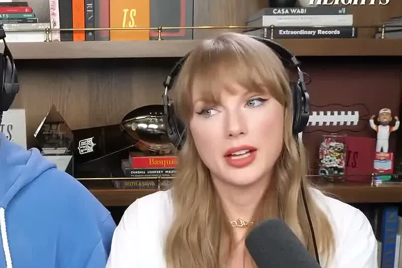 Taylor Swift wearing headphones and speaking into a microphone on the New Heights podcast set