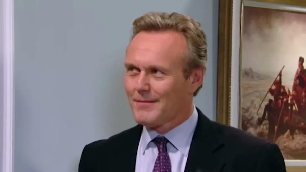 Anthony Head in a dark suit and purple tie smiling in an office set in Little Britain