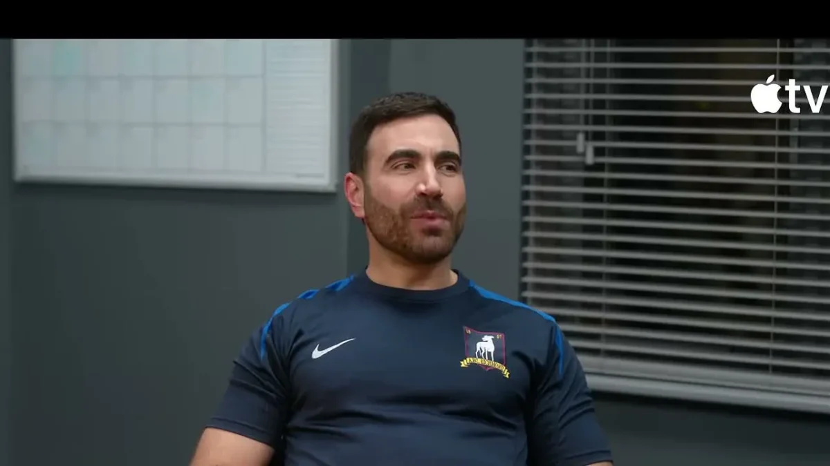 Brett Goldstein as Roy Kent in a navy AFC Richmond shirt sitting at a table in Ted Lasso