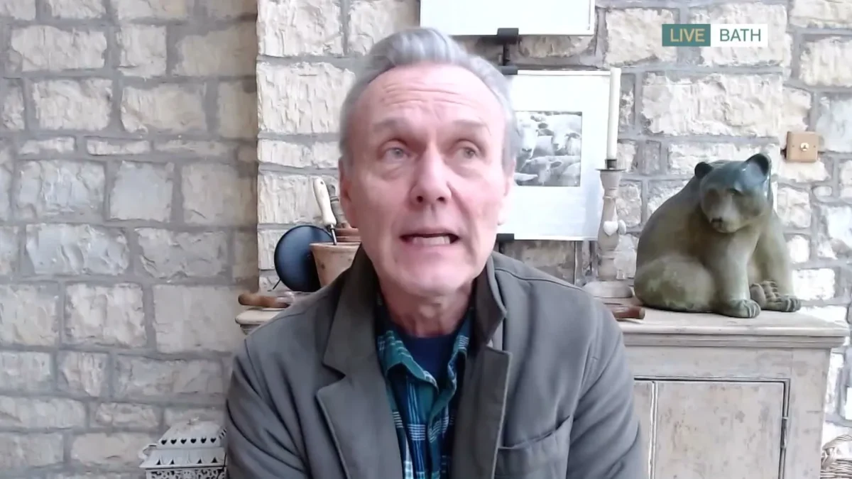 Anthony Head in a gray jacket and plaid shirt speaking on a video link from a stone-walled room
