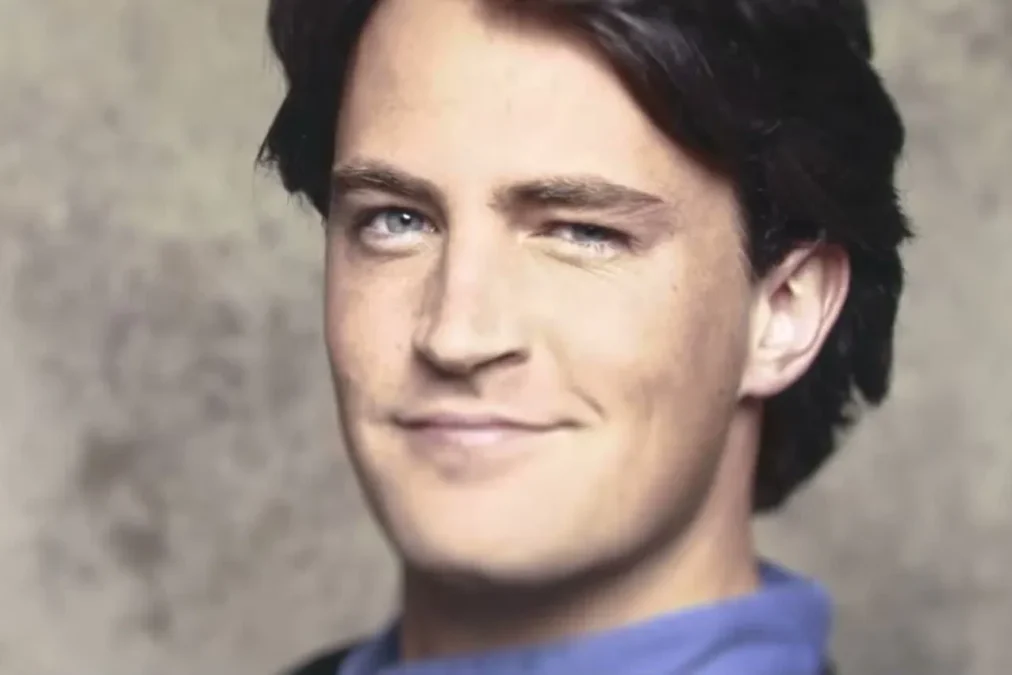 A young Matthew Perry with dark hair smiling slightly in a portrait against a gray backdrop