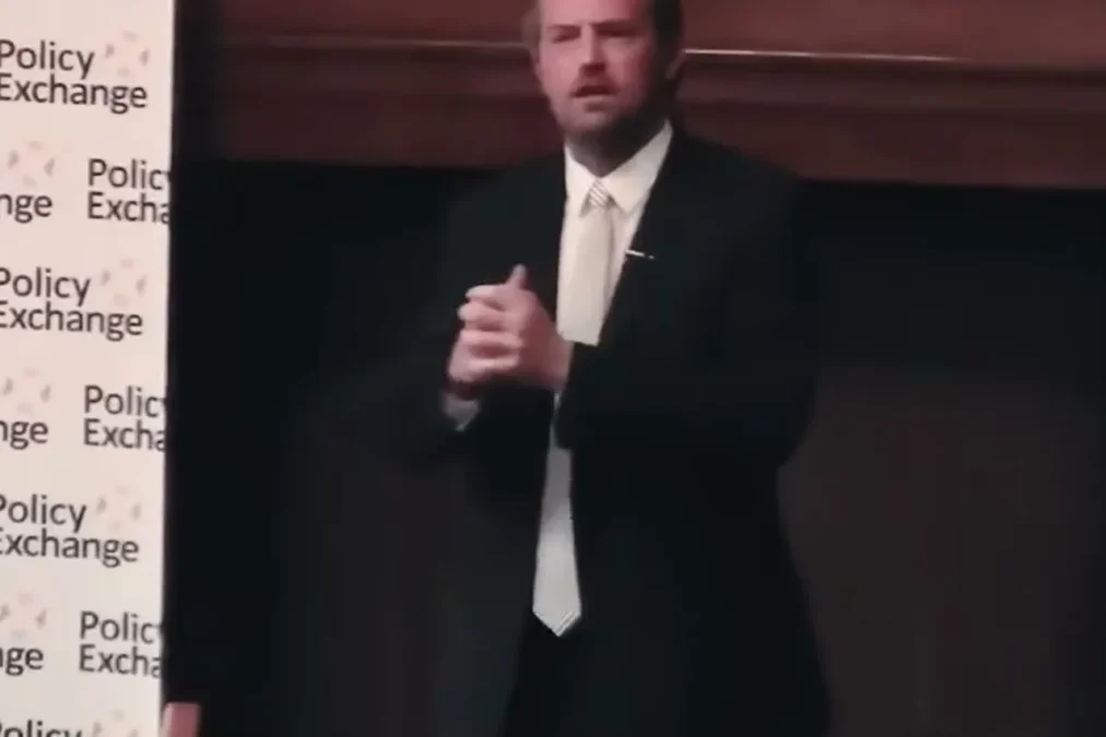 Matthew Perry in a dark suit and white tie speaking next to a backdrop printed with the words Policy Exchange