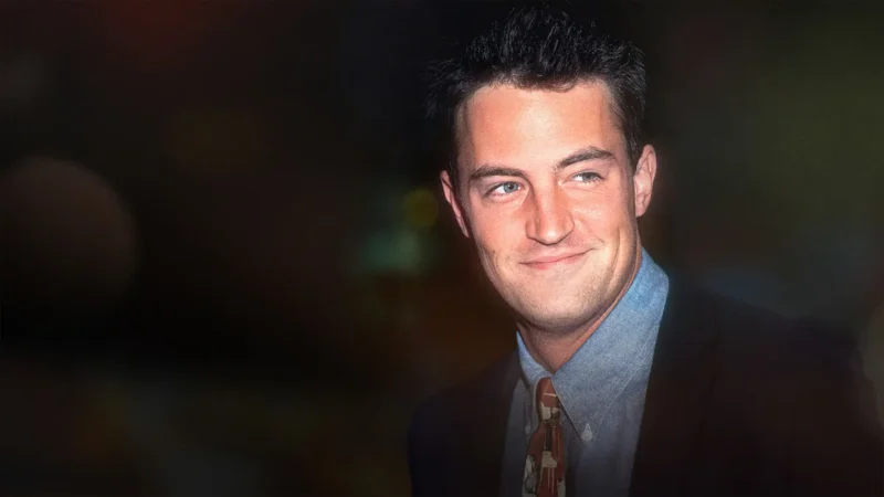 Matthew Perry smiling in a dark suit, blue shirt and patterned tie at an evening event
