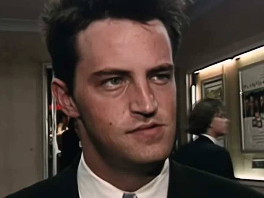 A young Matthew Perry in a black suit and white shirt in a hallway, looking to the side