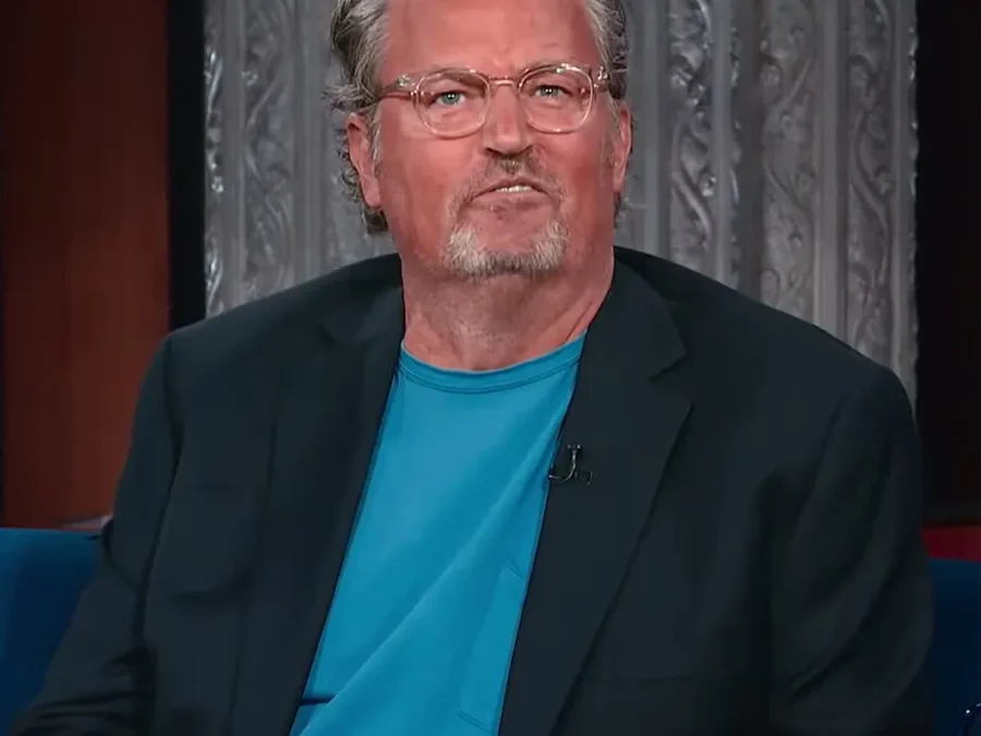 Matthew Perry with gray hair and glasses in a navy blazer and teal T-shirt, sitting on a talk show couch